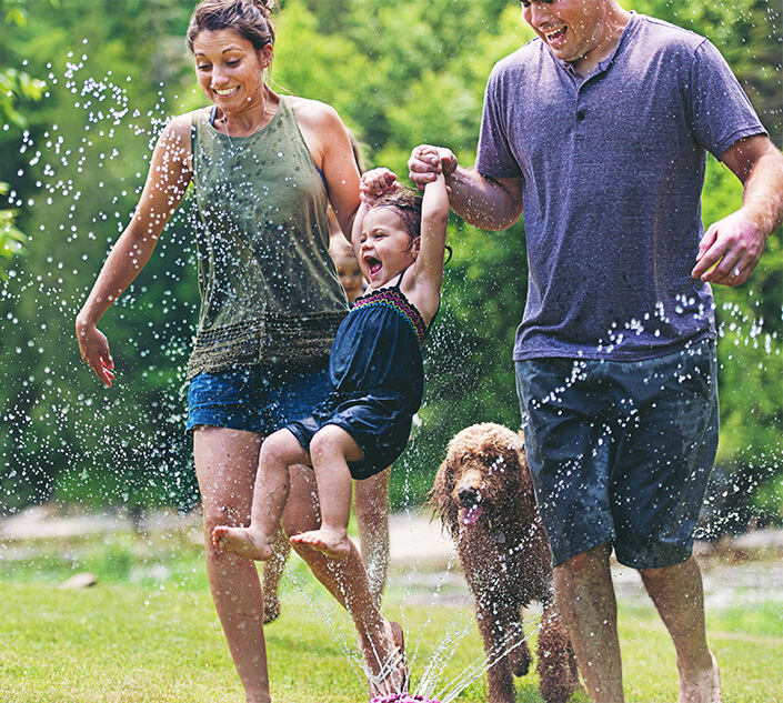 A family of three running through a sprinkler on the lawn